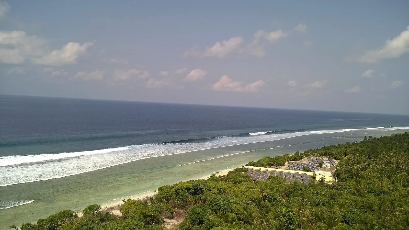 Elevated view over a green wooded shore, a pale fringing reef and surf breaking on the open sea